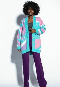 Sweter robiony na drutach w kolorach teal, różowym i fioletowym z geometrycznymi wzorami, z zapięciem na guziki i luźnym krojem, zestawiony z fioletowymi spodniami.