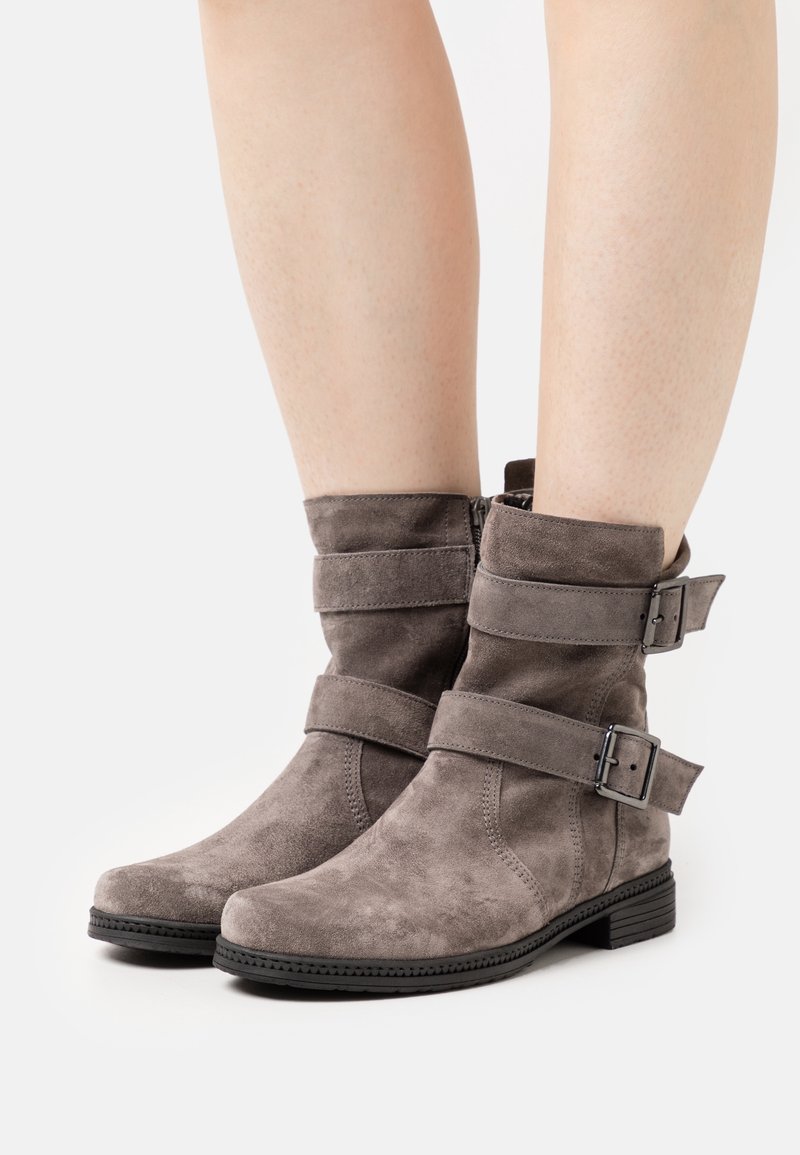 Bottes grises en daim montant à la cheville avec deux sangles réglables à boucle, portées sur des jambes nues, avec des semelles en caoutchouc noir basses et des détails de coutures.