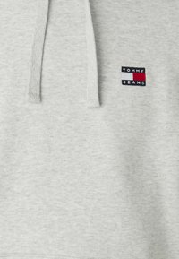 Sudadera gris claro con cordones, que presenta un pequeño logo bordado en azul marino, rojo y blanco. Textura suave y diseño casual.
