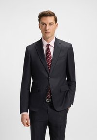 Mies, joka pukeutunut tummaan raitapukuun, vaaleanpunaiseen paitaan ja burgundinpunaraidalliseen solmioon, seisoo yhdellä kädellä taskussa valkoista taustaa vasten.