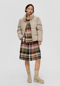 Beige gewatteerde jas, geruite jurk met groene, bruine, roze en witte patronen, en beige slouchy enkellaarsjes met dikke zolen.
