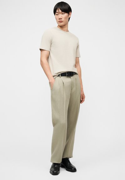 Άνδρας που φοράει μπεζ κοντομάνικο πουκάμισο, ανοιχτό μπεζ πλισέ παντελόνια, μαύρη ζώνη και μαύρα επίσημα παπούτσια, στέκεται με τα χέρια στις τσέπες.