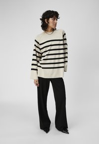 Maglione crema con strisce orizzontali nere, vestibilità ampia, texture a costine, abbinato a pantaloni neri a gamba larga e scarpe con punta. Collana in argento a catena.