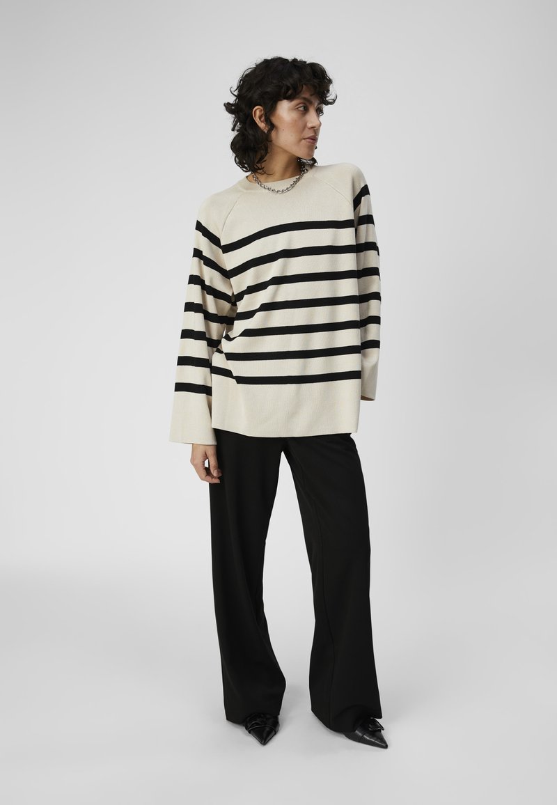 Cremefarbener Pullover mit schwarzen horizontalen Streifen, lockere Passform, gerippte Textur, kombiniert mit schwarzen weit geschnittenen Hosen und Schuhen mit spitzem Zehen. Silberne Kettenhalskette.