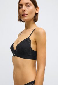 Crni čipkasti bralette s trokutastim oblikom košarica, podesivim naramenicama i čvrstim pojasom. Sadrži složene cvjetne detalje i mali metalni ukras.