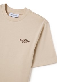 T-shirt en coton beige avec des manches courtes, un col rond et un design de texte imprimé marron indiquant "SUNDAY Brunch" sur la poitrine.