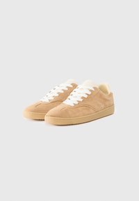 Lys brunsemulede sneakers med hvite snørebånd, rund tå og en teksturert gummisåle. Enkel design med minimale detaljer.