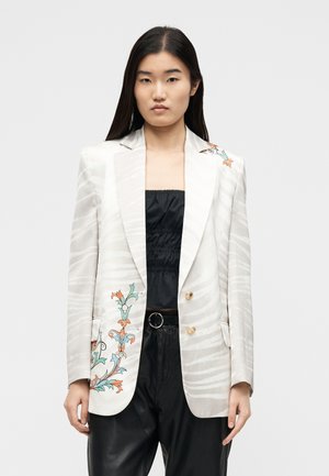 Ung kvinde med glat sort hår iført en hvid blazer med svage striber og blomsterbroderi, kombineret med sort rynket top og læderbukser.