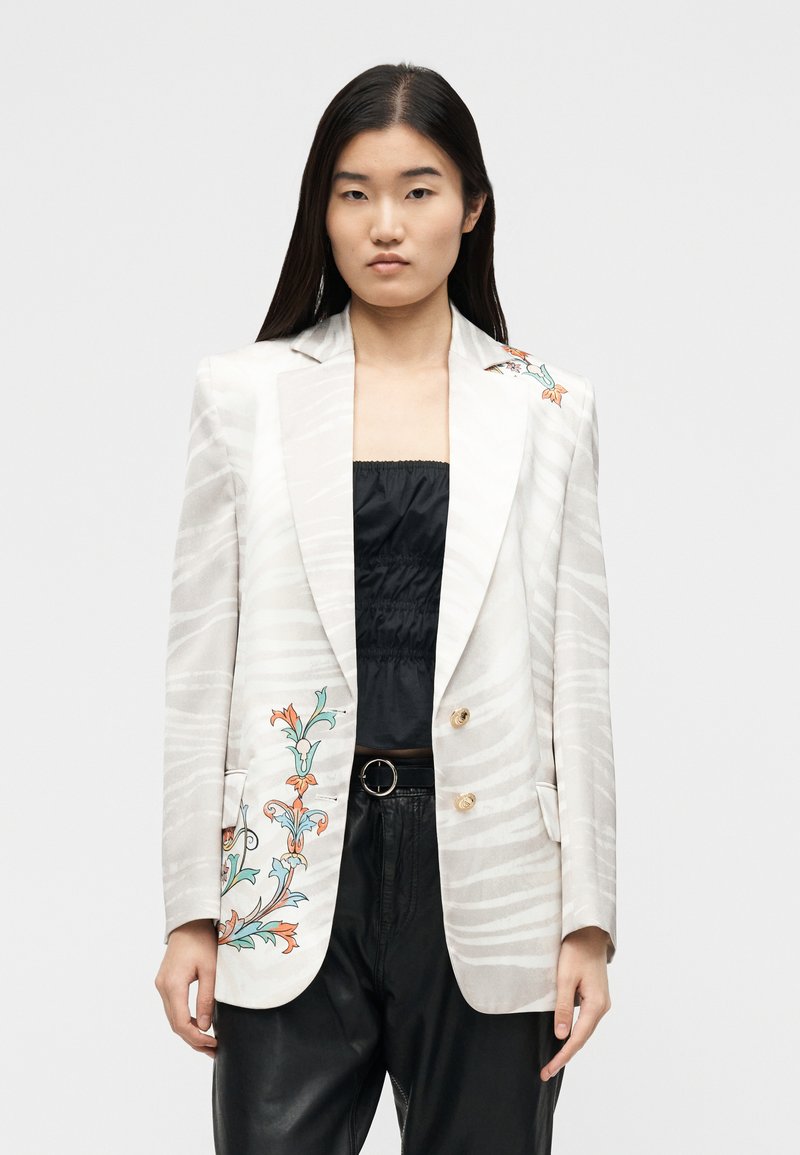 Jeune femme aux cheveux noirs et raides portant un blazer blanc à fines rayures et broderies florales, associé à un haut noir froncé et un pantalon en cuir.