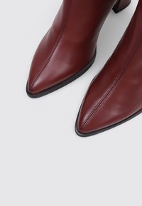 Botas de tobillo de cuero burdeos de tacón alto con punta afilada, que presentan costuras en contraste y una suela negra texturizada.