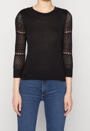 Schwarzer Strickpullover mit gemusterten Spitzen-3/4-Ärmeln, getragen zu blauen Jeans vor neutralem Hintergrund.