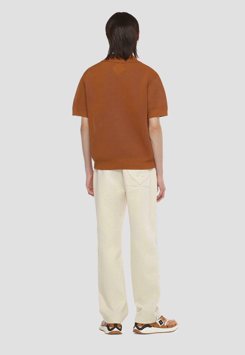 Braunes gestricktes kurzärmliges Hemd mit einem strukturierten Muster, kombiniert mit cremefarbenen Jogginghosen mit einem Logo auf der Rückentasche. Getragen mit modischen Schuhen.