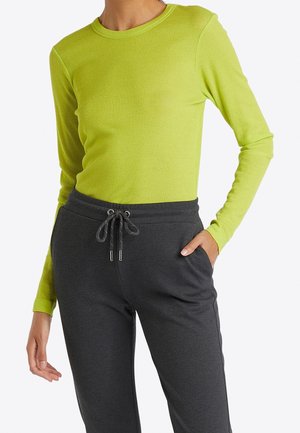 Frau trägt ein limettengrünes Langarmshirt und dunkelgraue Hose mit Kordelzug, eine Hand in der Hosentasche, vor weißem Hintergrund.