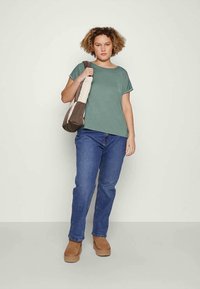 Top verde a maniche corte, jeans di denim blu, scarpe slip-on marroni, con una borsa a tracolla marrone e beige. Design semplice e rilassato.