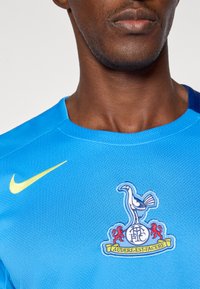 Gaiši zilā Nike krekls ar dzeltenu logo un kluba emblēmu, kurā attēlots putns un divi sarkani lauvas. Audumam ir teksturēta virsma.