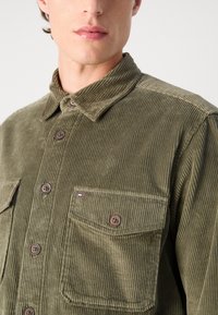 Groene corduroy overhemd met een button-down kraag, een borstzak, bruine knopen en subtiele textuurlijnen door het geheel.