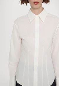 Femme portant une chemise blanche cintrée boutonnée avec un col classique, des manches longues, et des lèvres roses brillantes visibles, sur un fond clair uni.