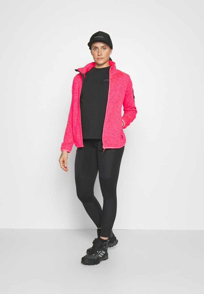 Donna con cappello nero, leggings neri e maglietta nera, indossa una giacca a zip rosa acceso aperta, in piedi contro uno sfondo bianco.
