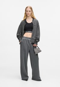 Sudadera gris con cremallera, top corto negro con logo, pantalones gris de pierna ancha en tejido suave, llevando un pequeño bolso de mano con un estampado.