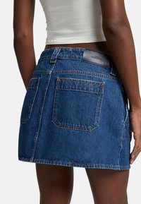 Jupe en denim mini bleu foncé, avec un design à cinq poches, des coutures visibles et un patch en cuir marqué sur la taille.