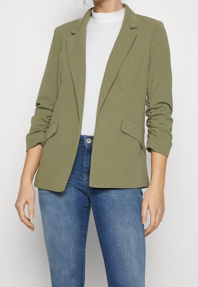 Personne portant un blazer vert olive avec des manches froncées, un haut blanc et un jean bleu sur un fond blanc uni.