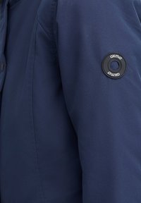 Veste bleu marine avec une texture lisse et mate. Dotée d'un logo noir circulaire sur la manche gauche, maintenu par un bouton pression.