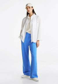 Biała koszula z długim rękawem na kremowym swetrze, w połączeniu z niebieskimi spodniami o szerokich nogawkach i białymi trampkami. Osoba ma także na sobie beżową czapkę.