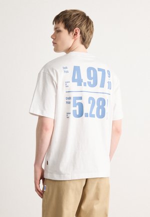 T-shirt bianca in cotone con grande testo stampato in blu sul retro. Il testo include prezzi in contante e con carta di credito con dettagli sulle tasse. Etichetta laterale visibile.