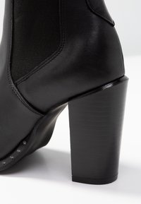Stivaletto in pelle nera con un pannello in tessuto testurizzato, tacco a blocco alto e dettagli in borchie argentate lungo il lato. Finitura liscia e forma strutturata.