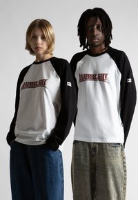 Due giovani adulti vestiti con magliette a maniche lunghe bianche e nere con il testo "WASTED", in piedi fianco a fianco contro uno sfondo semplice.