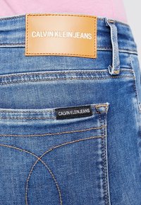 Ljusblå denimjeans med en ljusbrun läderetikett som står "CALVIN KLEIN JEANS" och en liten svart etikett på bakfickan. Kontraststickning synlig.