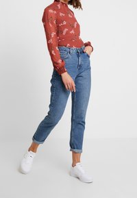 Kvinde iført en blomsterbluse i rustfarve med lange ærmer, højtaljede blå jeans med opsmøg og hvide sneakers, der går mod en ensfarvet hvid baggrund.