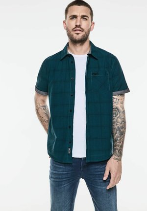 Kurzärmeliges grünkariertes Hemd mit schwarzen Knöpfen, Brusttasche und kontrastierenden grauen Ärmeln, getragen über einem weißen T-Shirt und blauen Jeans.