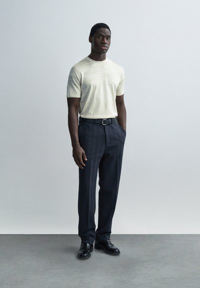 Ανοιχτόχρωμο, πλεκτό πουκάμισο με υφή συνδυασμένο με σκούρο παντελόνι σε ψαροκόκαλο, με ζώνη από μαύρο δέρμα, ολοκληρωμένο με μαύρα επίσημα παπούτσια.