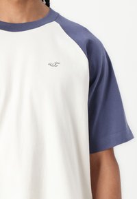 Raglan majica s bijelim tijelom i plavim rukavima. Ima mali logo na prsima. Izrađena je od mekog, prozračnog materijala.