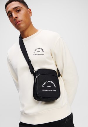 Borsa messenger nera con chiusura a zip e stampa del logo frontale. Tessuto liscio, forma rettangolare, tracolla regolabile. Indossata su una felpa bianca.