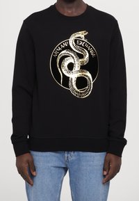 Svart crewneck sweatshirt med guldorms grafik och texten "Armani Exchange", bärs tillsammans med ljusblå jeans på en mörkhyad person.