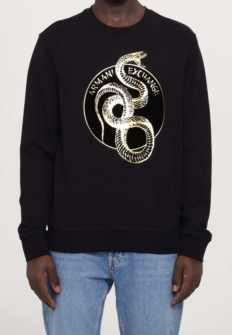 Svart crewneck sweatshirt med guldorms grafik och texten "Armani Exchange", bärs tillsammans med ljusblå jeans på en mörkhyad person.