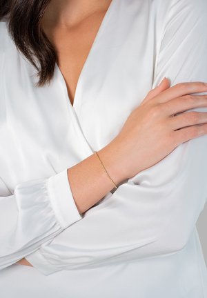 Gul armring med små runde detaljer, båret på et håndled, mod en baggrund af en hvid bluse med lange ærmer. Glat overflade.
