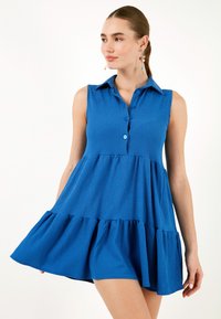 Rochie bleu fără mâneci, cu guler, având un decolteu în formă de cămașă, cu un încheiat pe față și fuste cu volane în straturi. Materialul texturat adaugă interes vizual.