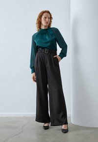 Femme aux cheveux bouclés roux portant un blouse en satin teal et un pantalon noir large taille haute avec des talons noirs, se tenant sur un sol en béton.