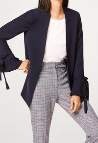 Nainen, jolla on tummansininen bleiseri hihnoilla, valkoinen paita ja korkeavyötäröiset harmaat ruudulliset housut, seisoo yksi käsi lanteella.