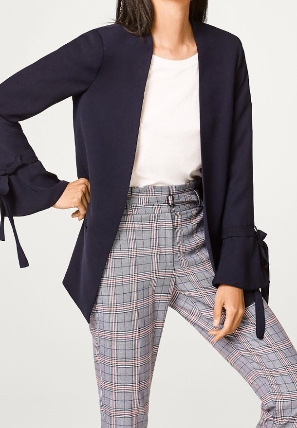 Nainen, jolla on tummansininen bleiseri hihnoilla, valkoinen paita ja korkeavyötäröiset harmaat ruudulliset housut, seisoo yksi käsi lanteella.