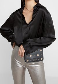 Kobieta w czarnej satynowej bluzce i srebrnych metalicznych spodniach, trzymająca czarną torebkę typu crossbody od Versace Jeans Couture z złotymi ćwiekami.