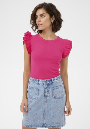 Frau trägt ein hellrosa Oberteil mit Rüschenärmeln und einen hellblauen Jeansrock mit pink bestickten Blumen, blickt zur Seite.