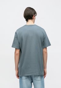 Νεαρός άνδρας με γυρισμένη πλάτη, φορώντας ένα ξεβαμμένο μπλε φαρδύ T-shirt και ανοιχτό μπλε τζιν τζιν, μπροστά σε ένα απλό φόντο.