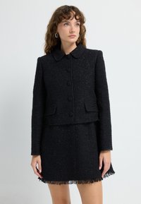 Sort tweedjakke med rund krave og fem knapper, parret med en matchende nederdel med frynsekant. Tekstureret stof hele vejen igennem.