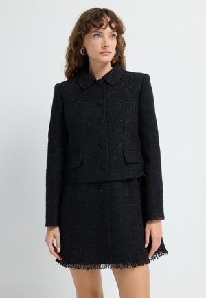 Sort tweedjakke med rund krave og fem knapper, parret med en matchende nederdel med frynsekant. Tekstureret stof hele vejen igennem.