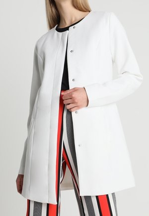 Femme portant un manteau blanc boutonné par-dessus un haut noir et un pantalon rayé avec des rayures verticales rouges, noires et blanches.