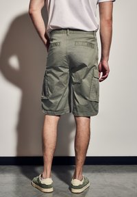 Zelené cargo šortky vyrobené z lehkého materiálu, s dvěma bočními kapsami, štíhlým designem a neformální délkou, doplněné zelenými teniskami.